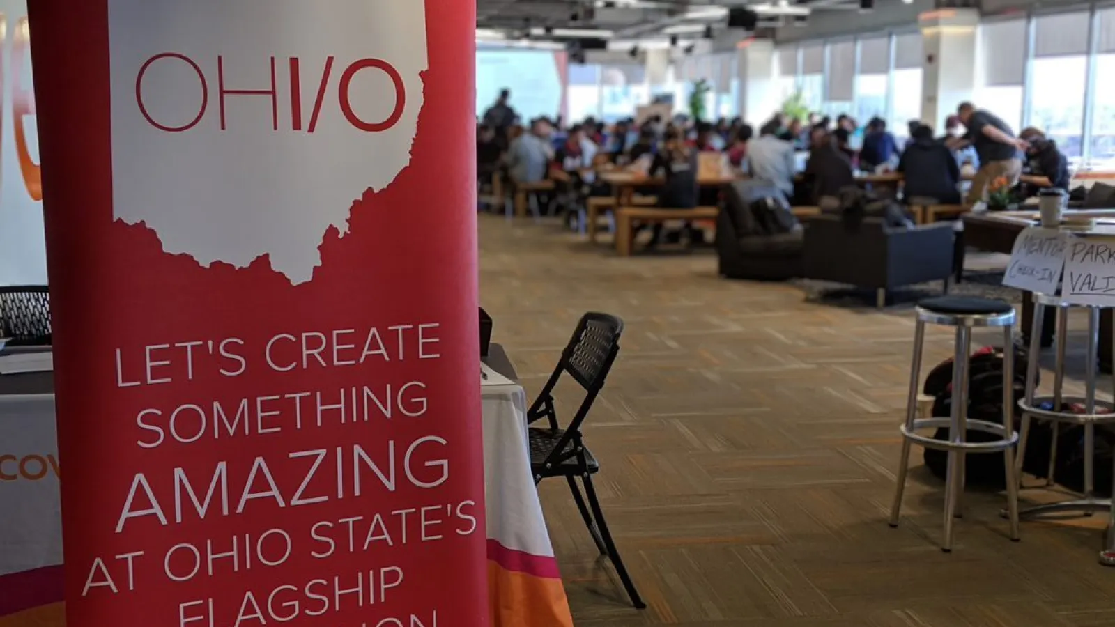 OHI/O banner in the foreground reads "Let's create something AMAZING at Ohio State's Flagship Hackathon", there are attendees working in the background