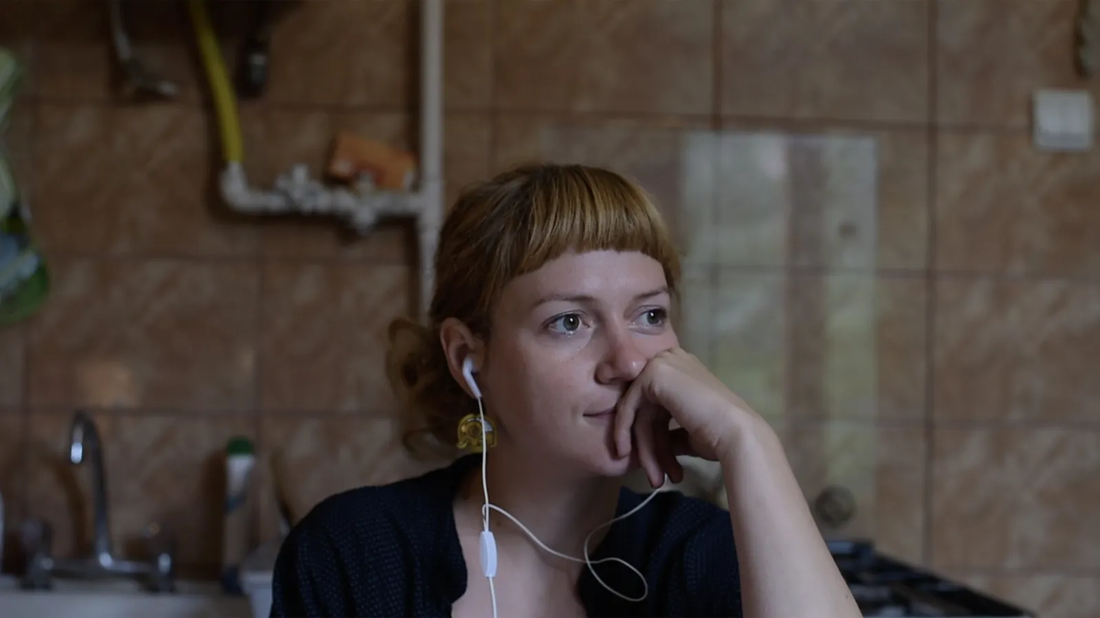 A women wearing earbuds with a saddened expression and thoughtful pose sits in a dark bathroom