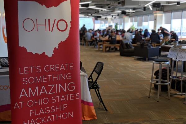 OHI/O banner in the foreground reads "Let's create something AMAZING at Ohio State's Flagship Hackathon", there are attendees working in the background