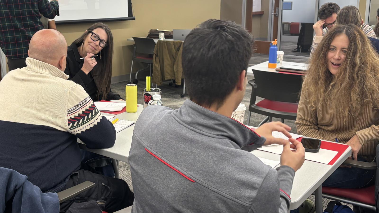Four people at a table, two with their backs to the camera and two facing the others with engaged looks on their faces